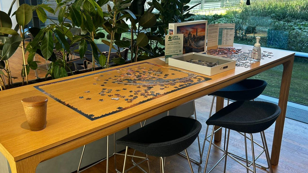 Long wooden table with a puzzle on top, surrounded by bar stools in a modern room with large windows.